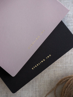A stack of B6 grid notebooks in matte dusty rose and black, each featuring the vertical foil-stamped brand name STERLING INK.