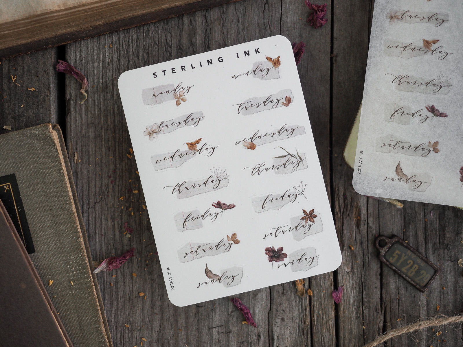 Decorative planner stickers labeling each weekday (Monday–Sunday) in rustic sepia script, accompanied by delicate printed illustrations of leaves and florals.